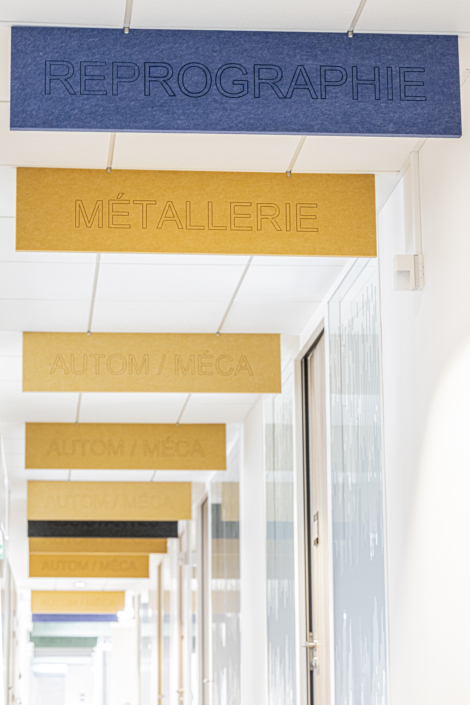 Pannelli blu e gialli, recanti le iscrizioni "REPROGRAPHIE", "MÉTALLERIE" e "AUTOM/MÉCA", sono sospesi al soffitto in uno spazio che ricorda una palestra acustica.
