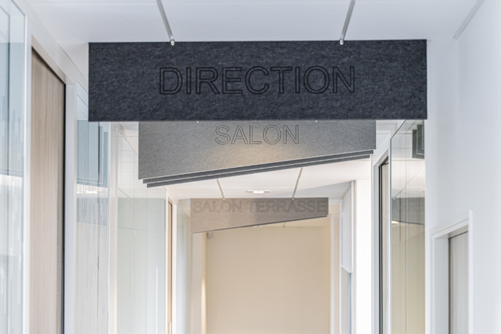Un corridoio ornato da pannelli a soffitto che indicano "MANAGEMENT", "LOUNGE" e "LOUNGE TERRACE" in ordine decrescente, che ricordano la chiara segnaletica di una palestra "acustica".