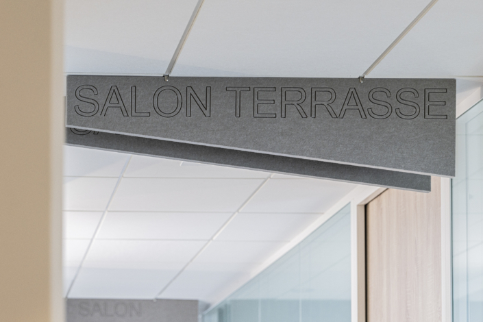 In un moderno corridoio d'ufficio con pareti in vetro, progettato secondo principi acustici spesso impiegati nelle palestre per ottimizzare l'isolamento acustico, un cartello grigio sospeso al soffitto reca la scritta "LOUNGE TERRACE".