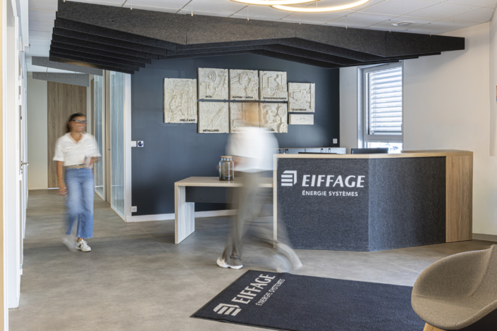 Due persone passeggiano nella hall di un moderno ufficio, dove i loghi di Eiffage sono visibili sul bancone della reception e sullo zerbino, mentre discutono delle recenti innovazioni in materia di acustica per palestre implementate negli ultimi progetti dell'azienda.