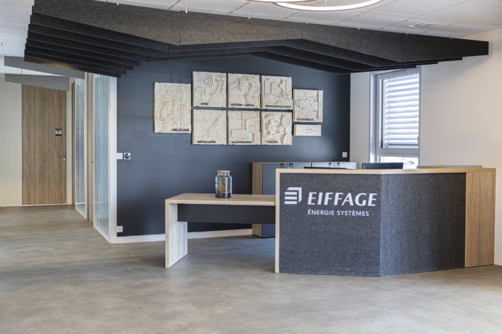Una moderna area reception per uffici, con bancone in legno, il logo Eiffage e un'opera d'arte appesa a una parete scura sullo sfondo, progettata per riflettere le linee pulite e le soluzioni acustiche ben studiate tipiche degli spazi contemporanei.