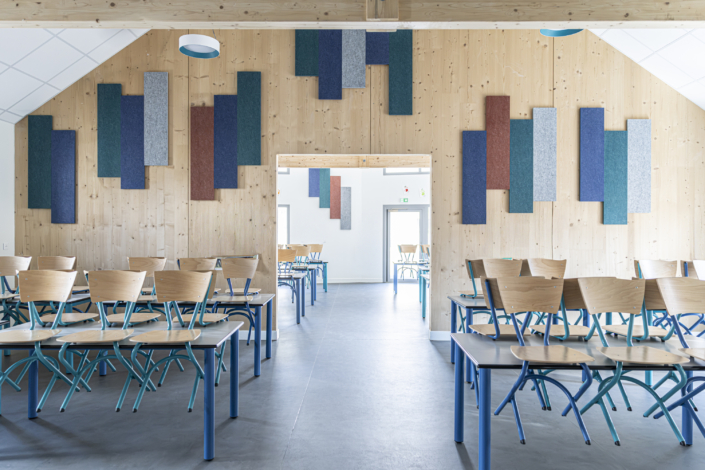 In un'aula luminosa, file di sedie e tavoli sono disposte in fila, con pannelli a parete colorati, dettagli in legno e un'acustica ispirata alle palestre per una migliore qualità del suono.