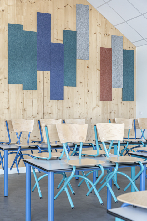 Un'aula vuota con banchi di legno, sedie blu e pannelli colorati fissati a una parete di legno, che ricordano gli impianti acustici dinamici delle palestre, in grado di migliorare sia lo stile che la qualità del suono degli spazi di apprendimento.