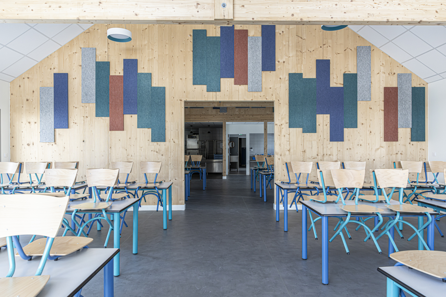 Aula moderna con pareti in legno, pannelli acustici colorati e file di sedie e tavoli vuoti.