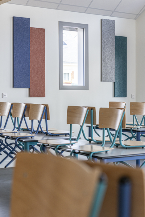 Aula vuota con sedie di legno appoggiate sui tavoli, una finestra e pannelli acustici colorati fissati alla parete.