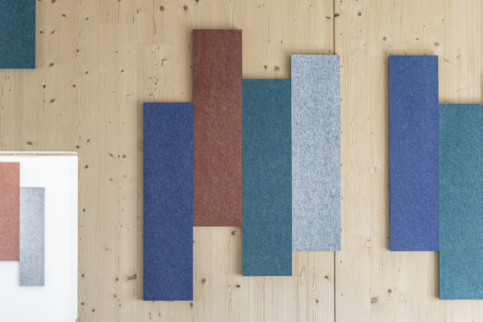 Pannelli rettangolari in tessuto blu, verde acqua, grigio e rosso, disposti su una parete di legno chiaro, creano un arredamento ispirato a una "palestra acustica".
