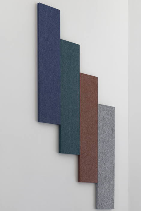 Quattro pannelli rettangolari nei colori blu, verde, rosso e grigio sono disposti diagonalmente su una parete bianca, creando una composizione accattivante ispirata all'acustica delle palestre.
