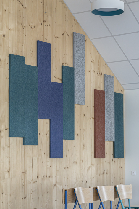 In una stanza moderna, pannelli rettangolari colorati, con un design ispirato alle palestre, sono fissati a una parete di legno sopra sedie blu e in legno.