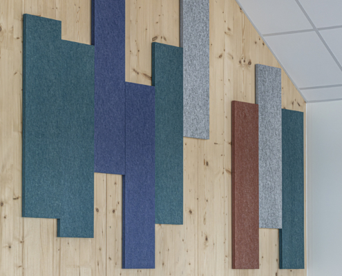 In una stanza moderna, pannelli rettangolari colorati, con un design ispirato alle palestre, sono fissati a una parete di legno sopra sedie blu e in legno.