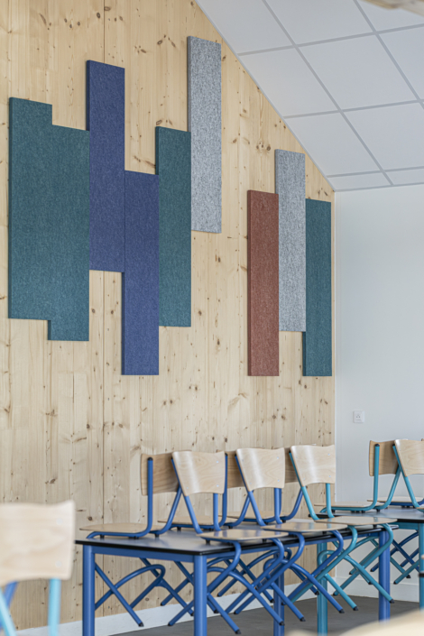 Aula moderna caratterizzata da una parete in legno, pannelli verticali colorati progettati per fornire un isolamento acustico di qualità "da palestra" e sedie in legno impilate su banchi in metallo blu.