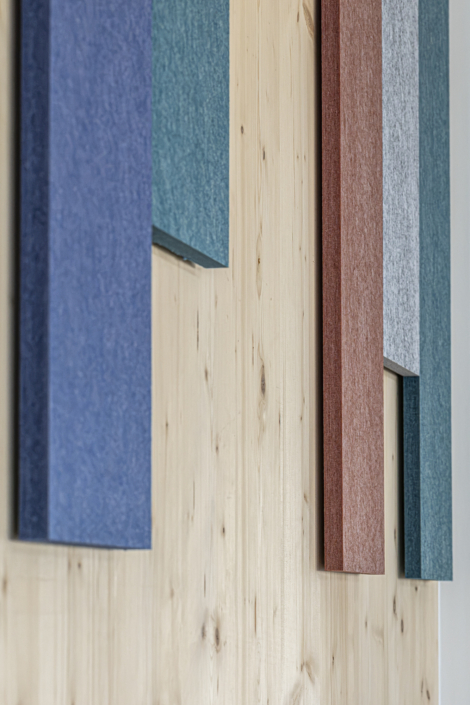 Pannelli acustici rettangolari di colore blu, verde, rosso e grigio sono fissati verticalmente a una parete di legno nella palestra.