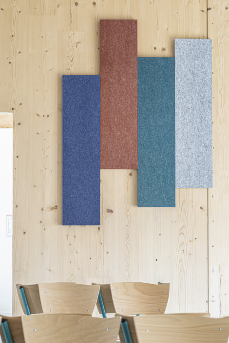 Quattro pannelli di tessuto colorato sono sospesi verticalmente su una parete di legno chiaro, sopra alcune sedie di legno, fornendo una soluzione acustica adatta alle palestre che migliora sia la qualità del suono che l'estetica dello spazio.
