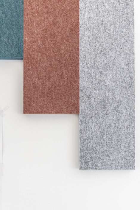 Tre pannelli rettangolari in feltro, nei colori verde acqua, ruggine e grigio, sono appesi a una parete bianca, offrendo un tocco di design acustico moderno che ricorda un'elegante palestra.