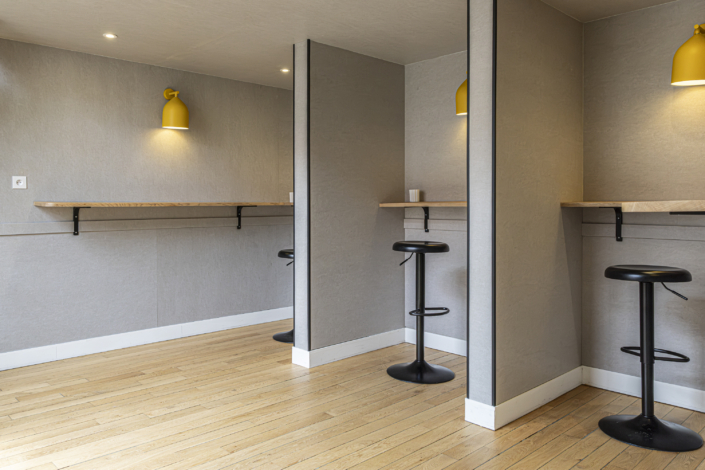 Uno spazio di lavoro minimalista con sgabelli alti, banconi in legno, pareti grigie, applique gialle ed elementi di design ispirati alle palestre, pensati per migliorare l'isolamento acustico.