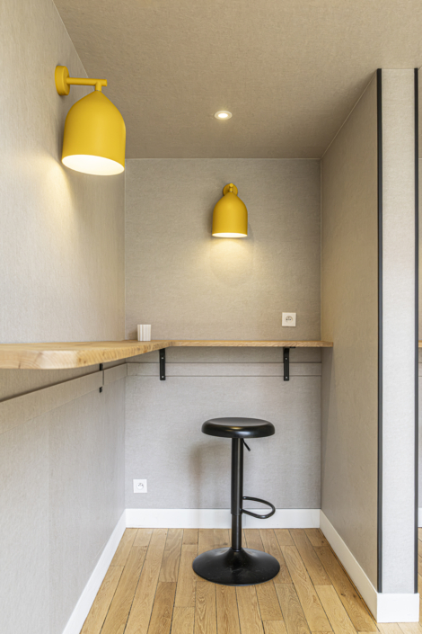Uno spazio di lavoro minimalista composto da uno sgabello nero, una scrivania in legno, due applique gialle e pareti grigio chiaro, ispirato al design acustico delle palestre per una migliore qualità del suono.