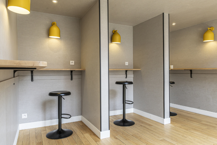 Tre postazioni di lavoro dotate di sgabelli alti, banconi in legno e applique gialle, disposte in un ambiente moderno e minimalista, offrono un'oasi di pace che ricorda una palestra acustica, coniugando funzionalità e design sereno.