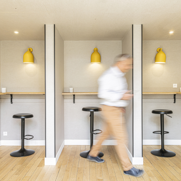 Un homme dont la silhouette est floue passe devant trois postes de travail équipés de tabourets noirs et de lampes jaunes, dans une pièce moderne conçue selon les principes de l'acoustique des gymnases.