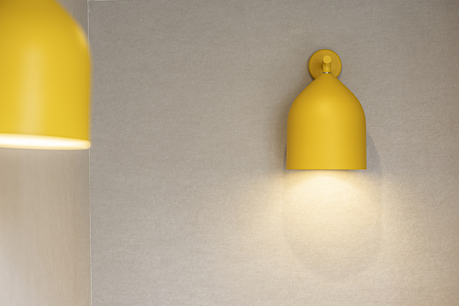 Una lampada da parete gialla è accesa e illumina una semplice parete beige che ricorda le linee pulite che si trovano spesso nelle palestre dotate di impianto acustico, mentre un'altra lampada è parzialmente visibile sulla sinistra.