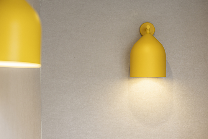 Una lampada da parete gialla è accesa e illumina una semplice parete beige che ricorda le linee pulite che si trovano spesso nelle palestre dotate di impianto acustico, mentre un'altra lampada è parzialmente visibile sulla sinistra.