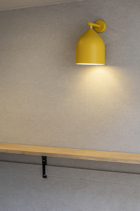 Una lampada da parete gialla illumina una mensola in legno chiaro addossata a una parete beige, accentuando l'atmosfera serena che ricorda quella di una palestra acustica.