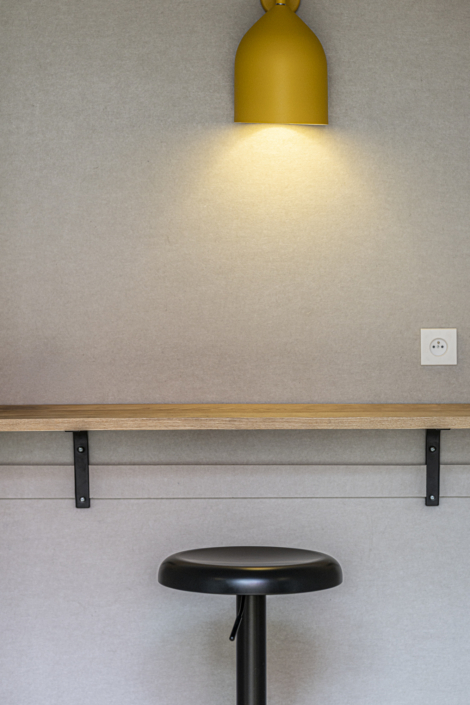 Sotto un bancone di legno è posizionato uno sgabello nero, sul quale si trovano una lampada gialla e una presa elettrica, il tutto di fronte a una parete beige, evocando il fascino minimalista di una palestra acustica.