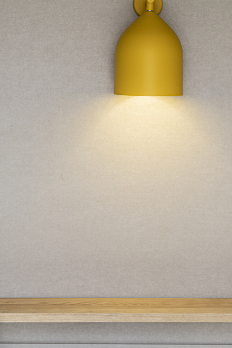 Una lampada da parete gialla illumina una parete beige vuota sopra una semplice mensola di legno, evocando l'atmosfera silenziosa di una palestra acustica.