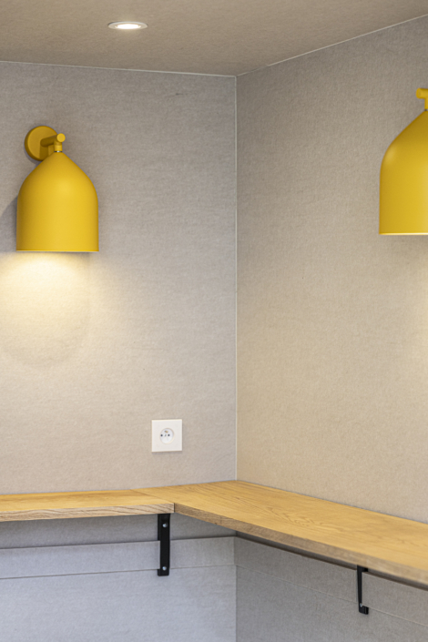 Due applique gialle illuminano un angolo con pareti beige, sormontato da una mensola in legno, creando un'atmosfera calda che ricorda quella di una palestra acustica.